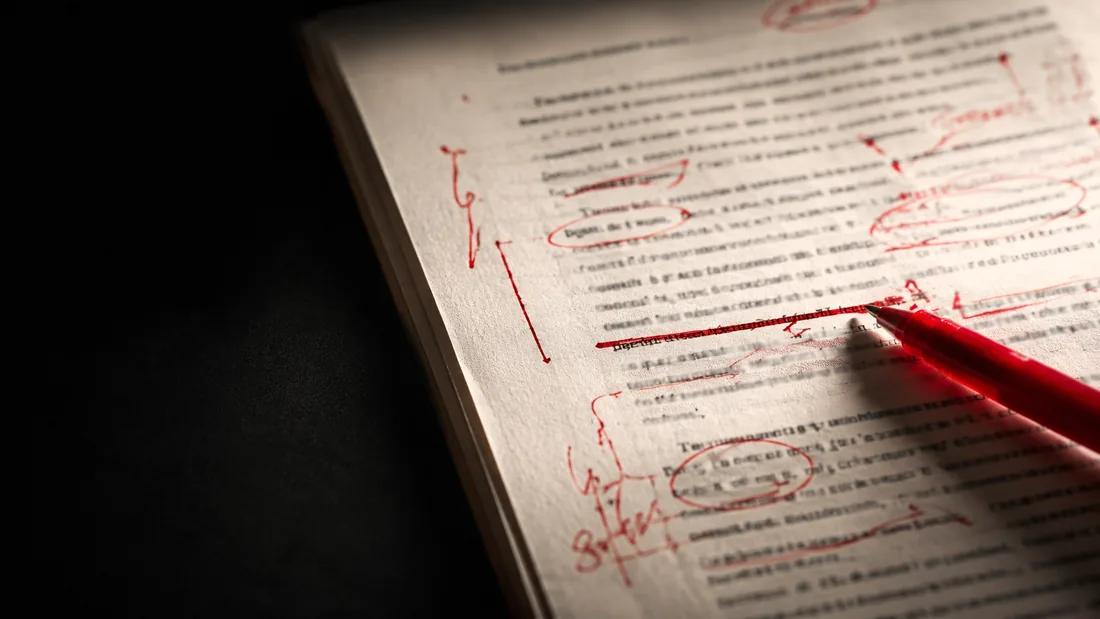 A printed manuscript covered in red-pen editing marks, corrections, strike-throughs, and circled errors under a desk lamp